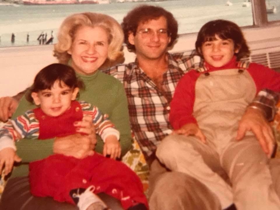 A sunlit early-1980s family snapshot by the water in New London, Connecticut: a toddler in red on the left, an older woman with blonde hair in a green sweater, a young Robert Rand in a plaid shirt and glasses, and a child in tan overalls on the right.